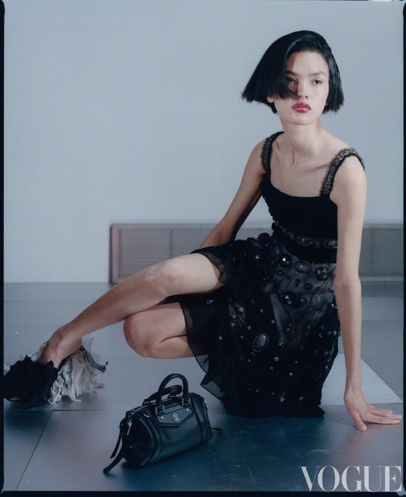 Fashion photography features a woman in an embellished black dress kneeling indoors next to a black handbag, with a neutral grey background.