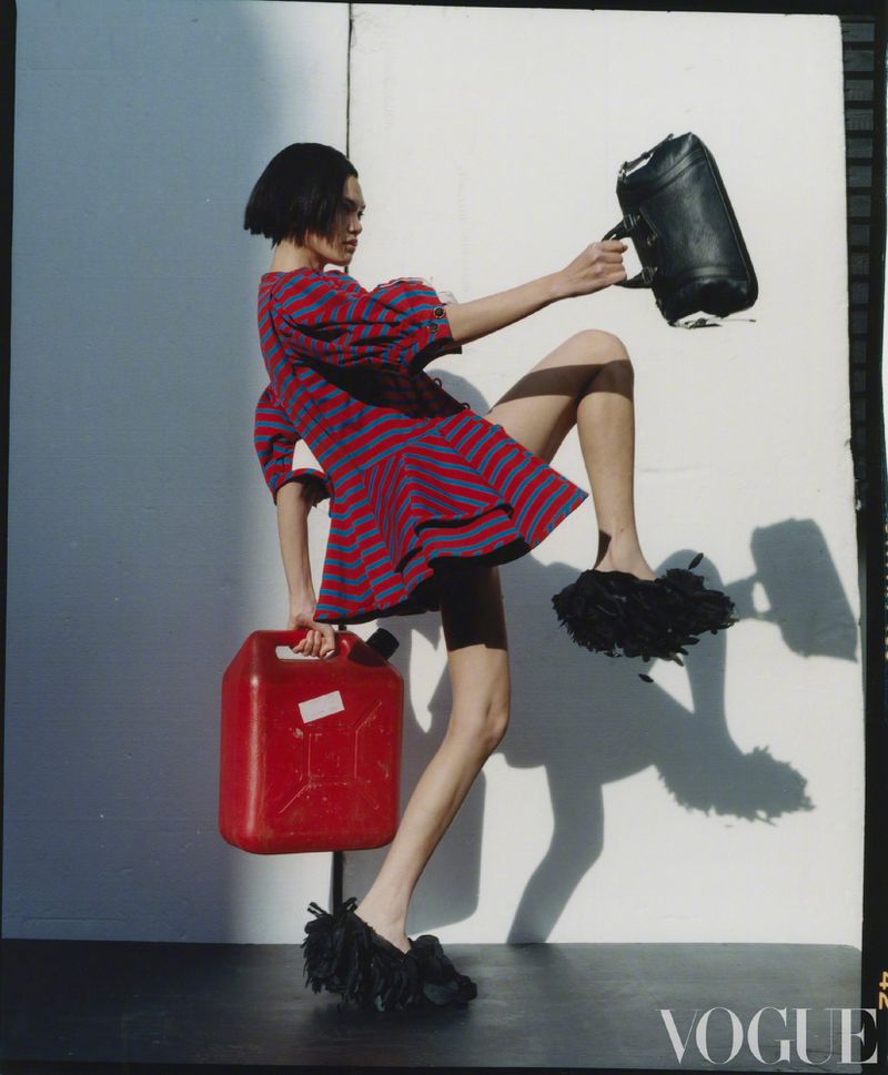 A fashionable woman in a striped dress poses with a red jerrycan and a black leather bag against a minimalist white backdrop.