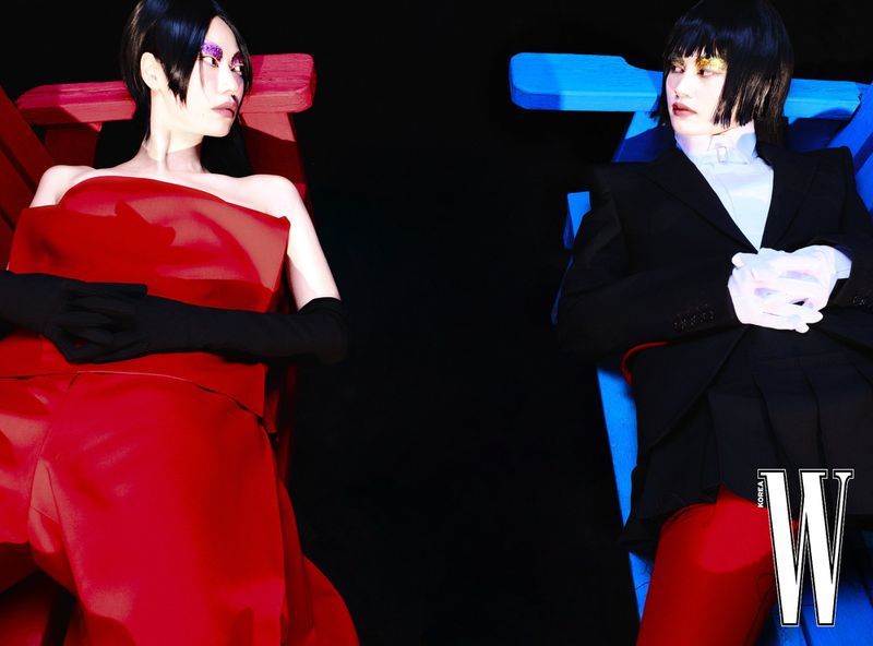Two models in striking red and black and white outfits pose in a photo studio against a black background with dramatic lighting.
