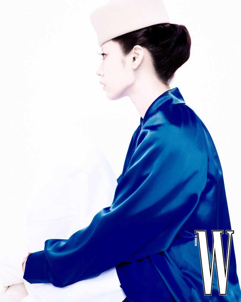 Fashion portrait of an adult woman with an updo wearing a blue satin jacket and cream-colored hat against a bright white background.