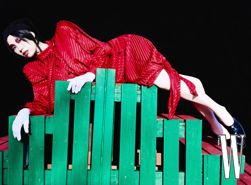 Fashionable person lies on a green wooden structure wearing a red striped outfit, white gloves and black flats in a studio setting.