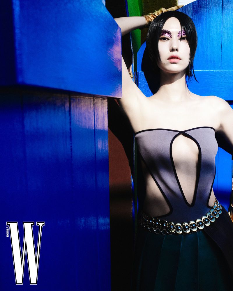 Woman with glitter makeup poses in front of blue wall wearing gray strapless top and long dark skirt for fashion editorial.