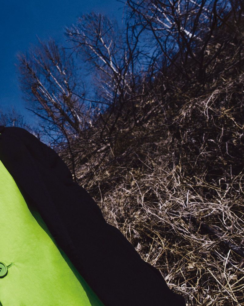 Low angle shot of a person wearing a bright green coat against a dry brushwood hillside and blue sky.