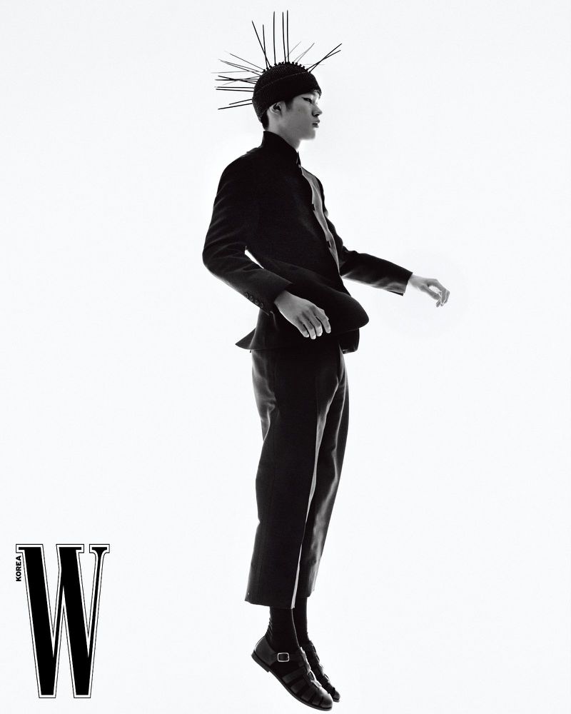 Monochrome fashion shot of an Asian male model in a dark suit and headdress with spikes, floating against a bright white background in a studio.