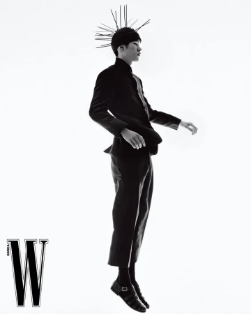 Monochrome fashion shot of an Asian male model in a dark suit and headdress with spikes, floating against a bright white background in a studio.
