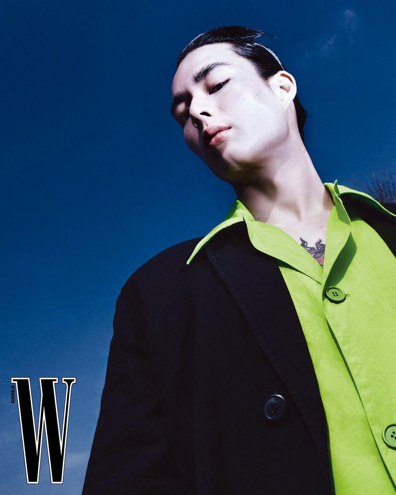 Portrait of an Asian man wearing a green shirt and black jacket against a blue sky, shot at a low angle, showcasing fashion.