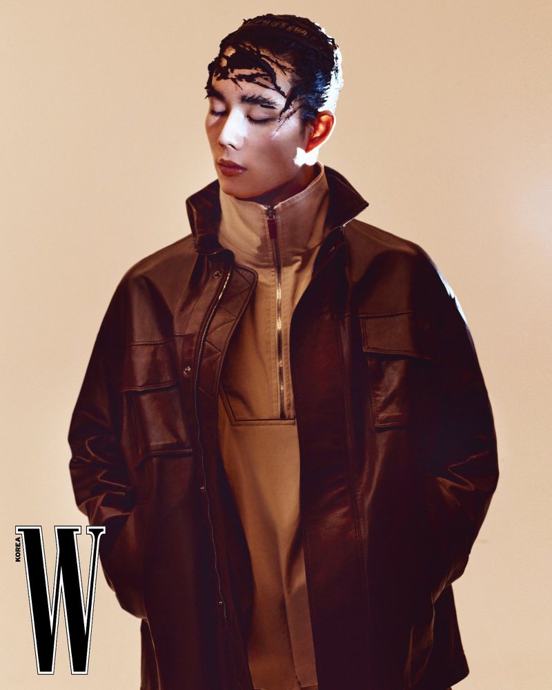 Fashion editorial shot: a young adult with unique headwear poses in a brown leather jacket against a neutral background in a studio setting.