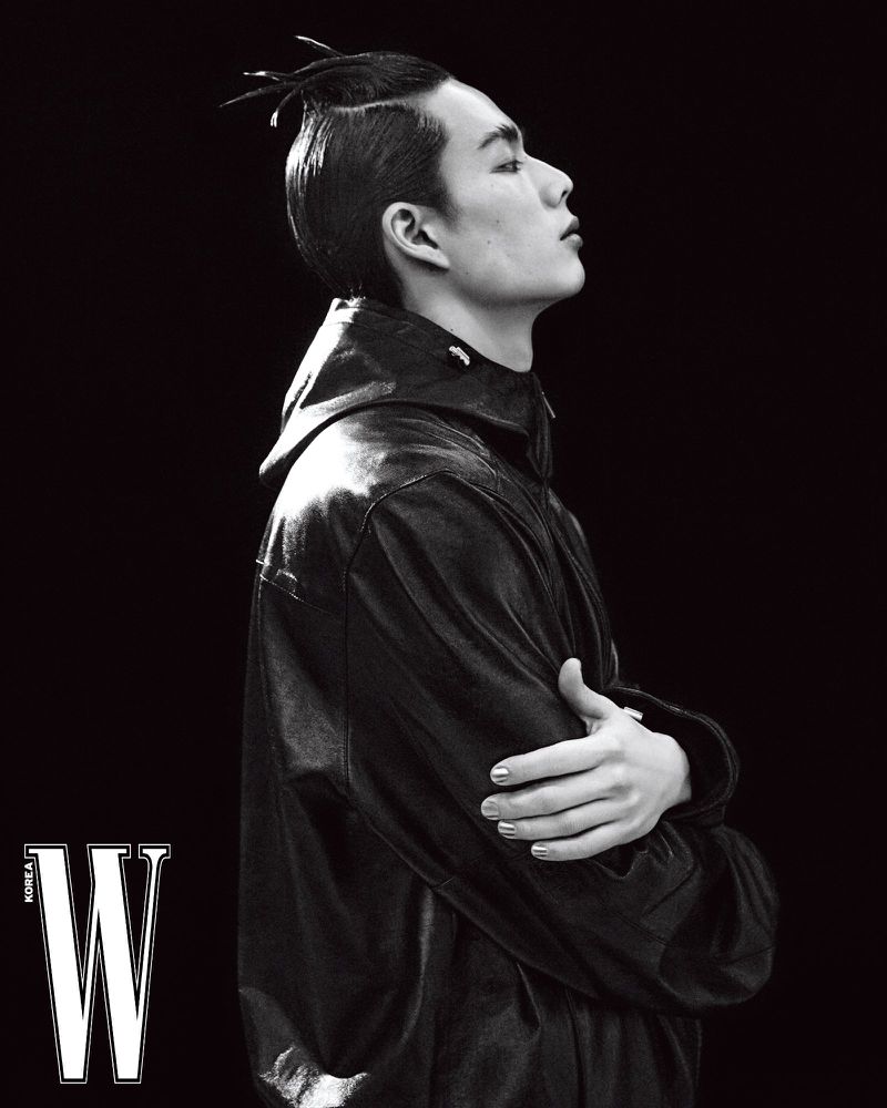 Black and white portrait of an adult male with unique hairstyle wearing a black jacket against a dark backdrop, fashion editorial.