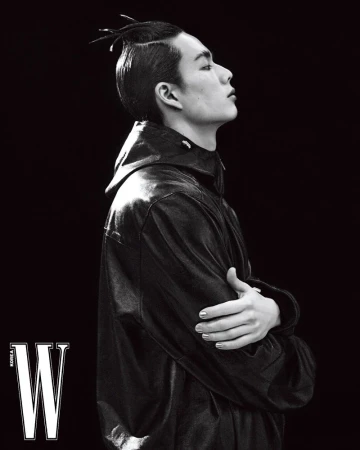 Black and white portrait of an adult male with unique hairstyle wearing a black jacket against a dark backdrop, fashion editorial.