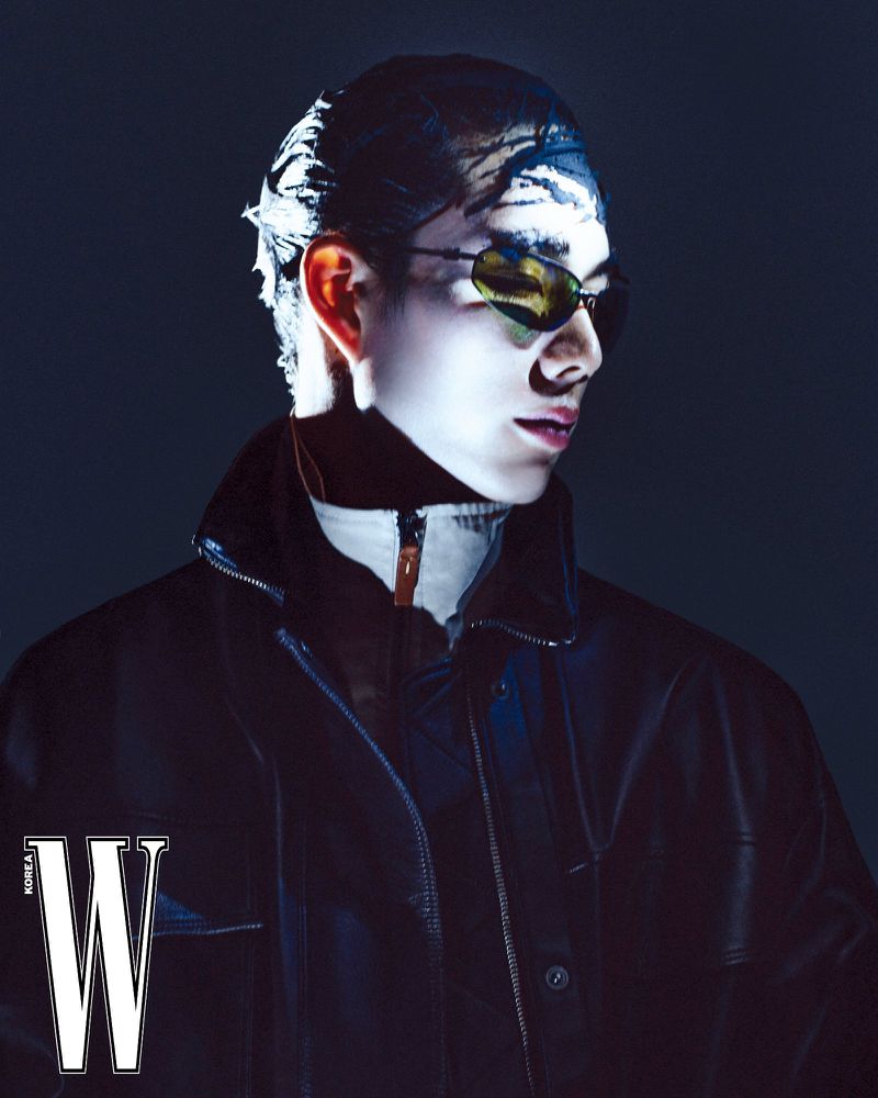 Portrait of an adult male in sunglasses and a dark jacket with a turtleneck collar, posing in a photo studio with 'W Korea' magazine logo.
