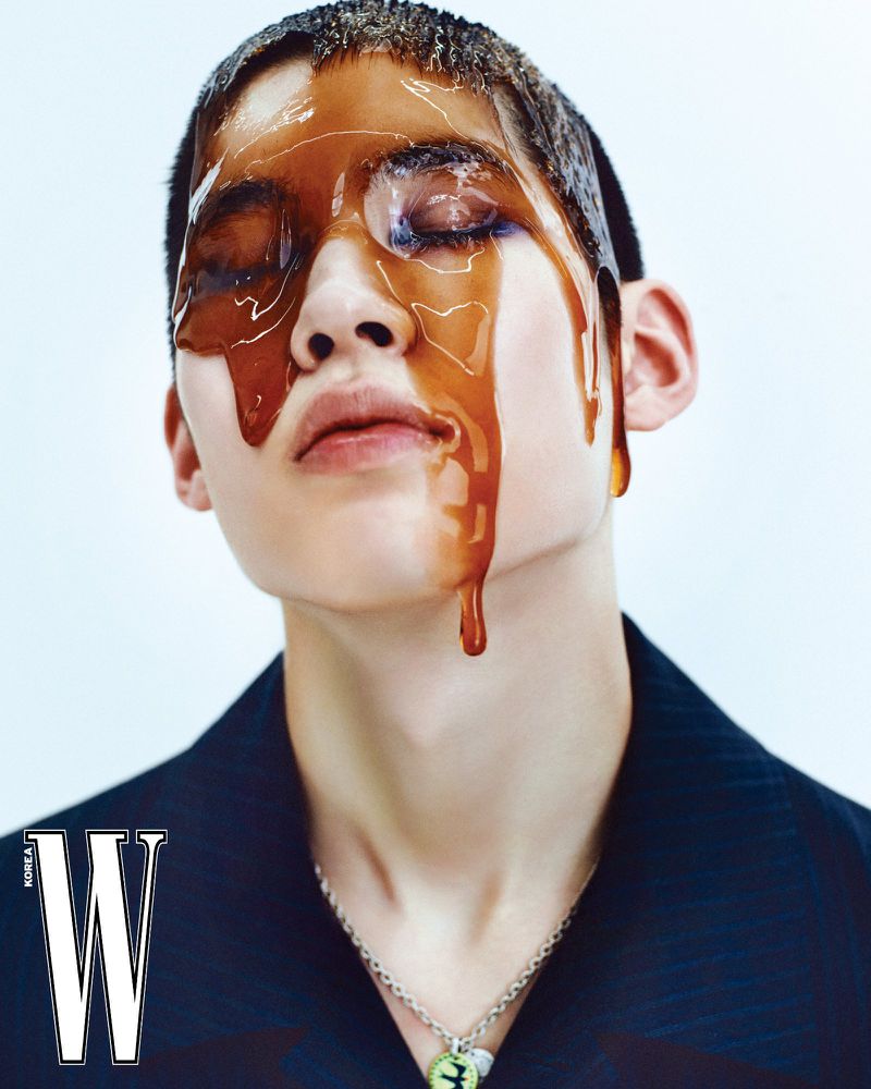Fashion shot of a model with glossy liquid dripping down their face, wearing a dark jacket and necklace, against a gradient backdrop.