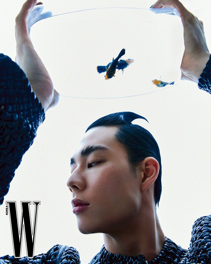 Editorial portrait of a slick-haired Asian male model in a woven top, holding a bowl of goldfish in a bright, high-key indoor fashion shot.