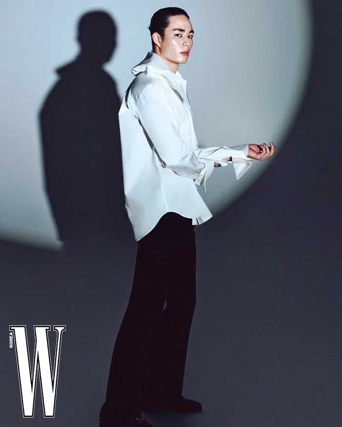 Asian male model wearing a white button-down shirt and black trousers standing against a grey wall with a circular spotlight projection.