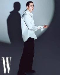 Asian male model wearing a white button-down shirt and black trousers standing against a grey wall with a circular spotlight projection.