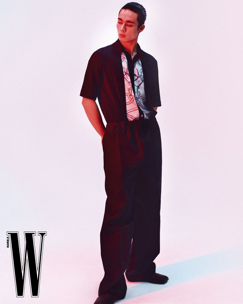 Model wearing a black short-sleeve shirt with a patterned scarf detail and dark trousers against a white backdrop.