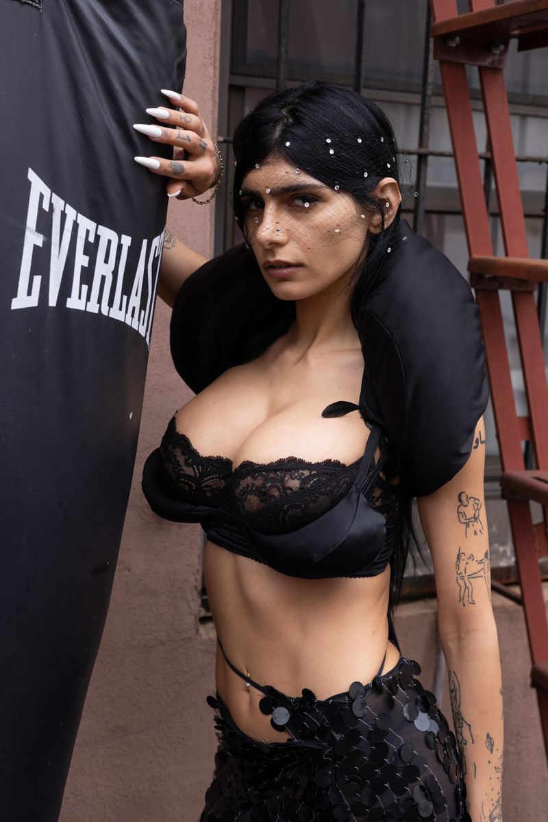 Brunette woman in a black bra poses beside an Everlast punching bag in front of a brick building with fire escape.