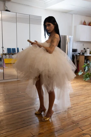A young adult female with dark hair is indoors wearing a large white tulle dress with gold heels, holding a cellphone.