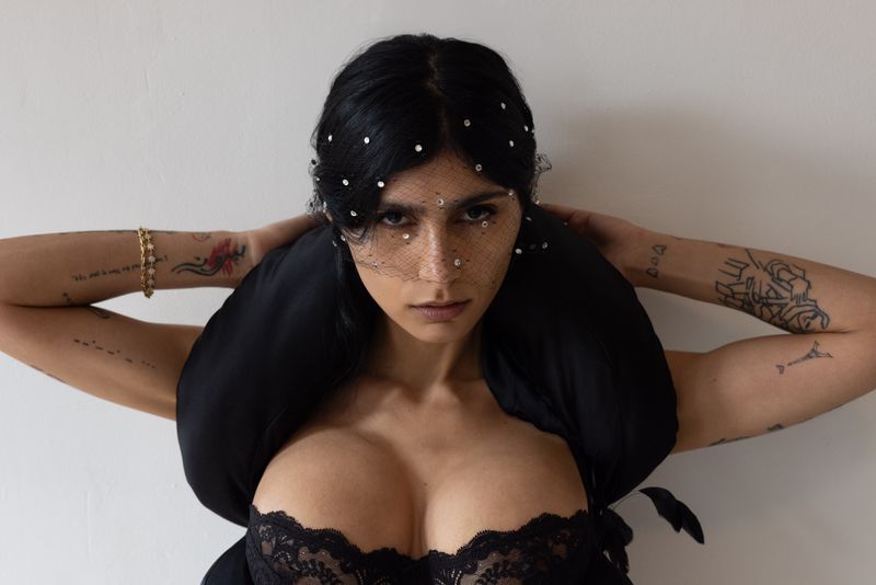 Close-up shot of a woman with dark hair wearing black lace and a beaded veil, posing in front of a plain white wall.