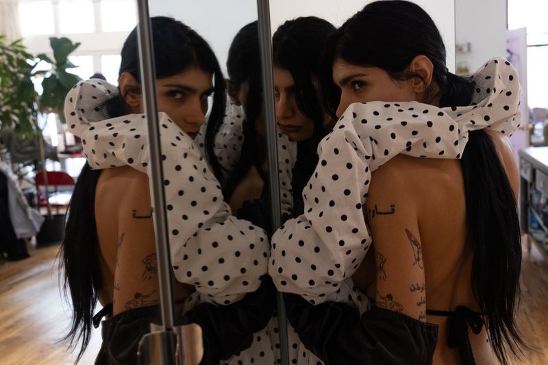 Woman with long black hair poses in a white polka dot blouse in front of a mirror reflecting multiple angles of her face and body.