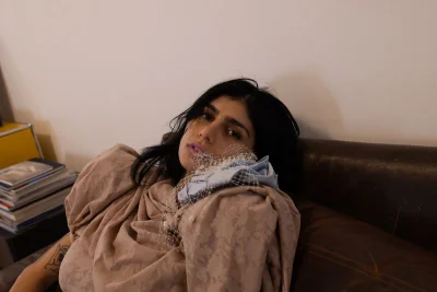 Mia Khalifa resting on a brown leather couch wearing a draped beige patterned garment with a veiled accessory.