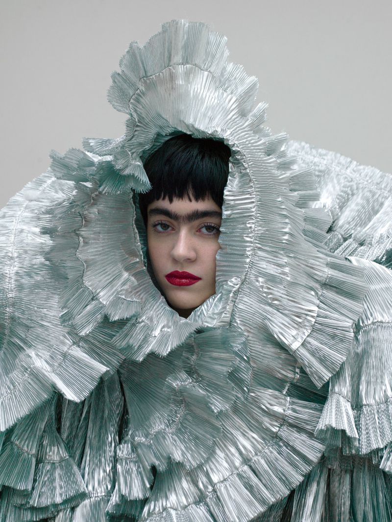 Woman with dark hair and bright red lips is posing wearing a large, ruffled silver-blue garment against a grey background in a studio shot.