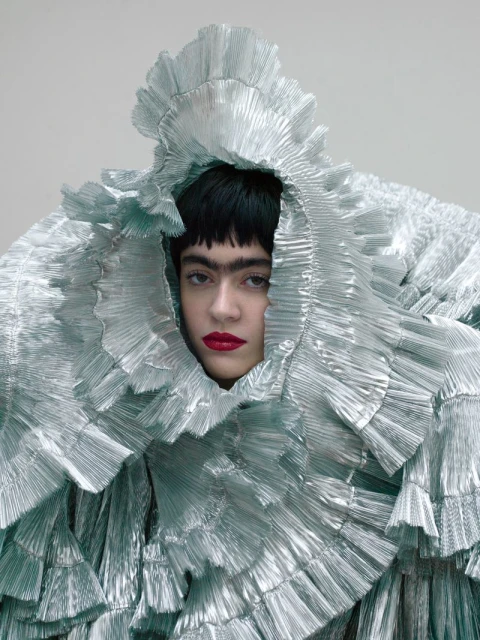Woman with dark hair and bright red lips is posing wearing a large, ruffled silver-blue garment against a grey background in a studio shot.