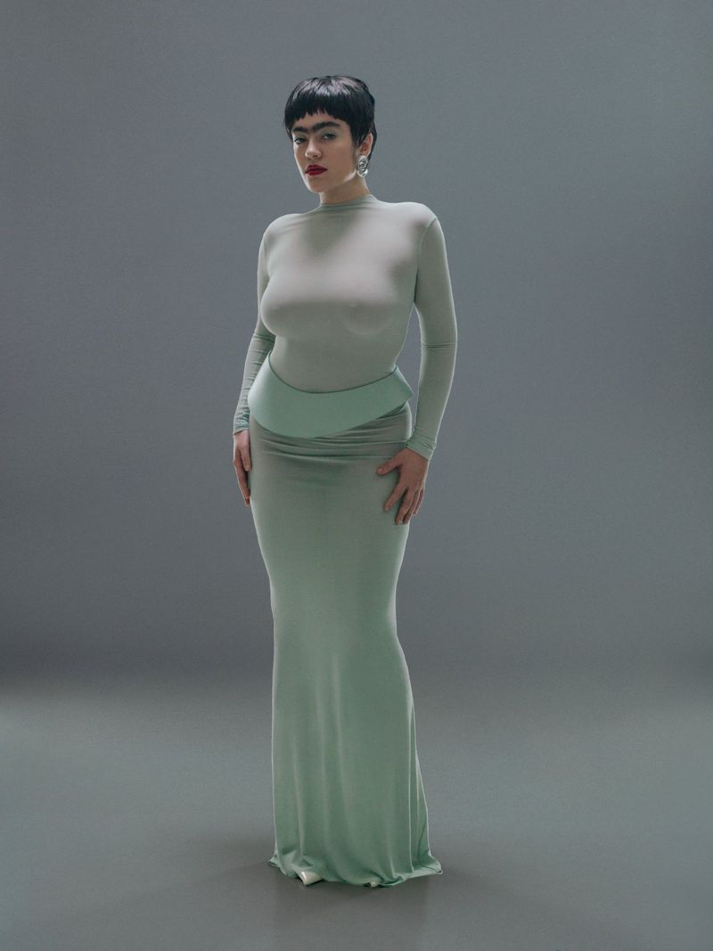 Full-length portrait of a woman with dark hair in a light green, full-sleeved top and a matching long skirt against a grey backdrop.
