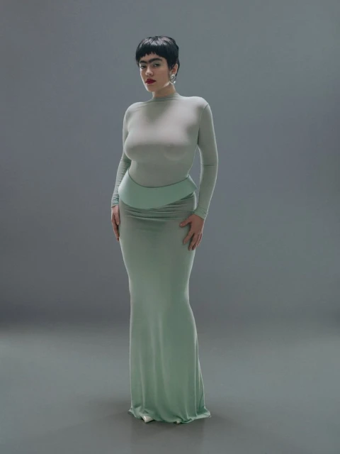 Full-length portrait of a woman with dark hair in a light green, full-sleeved top and a matching long skirt against a grey backdrop.