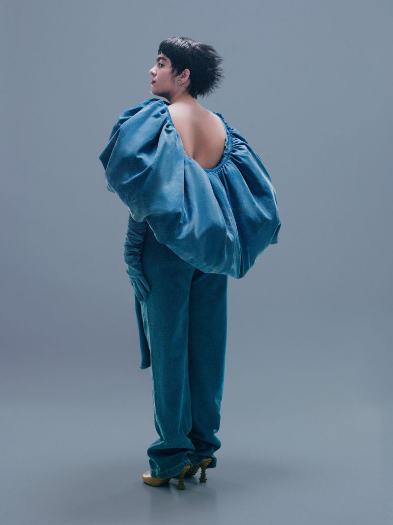 Adult female poses in an open back teal pantsuit with gloves and pointed-toe heels against a neutral grey studio backdrop in full shot.