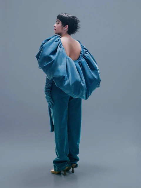 Adult female poses in an open back teal pantsuit with gloves and pointed-toe heels against a neutral grey studio backdrop in full shot.