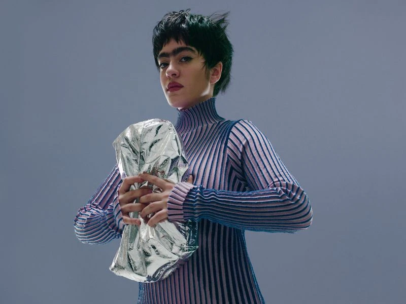 Woman in ribbed top holds a reflective silver object in a studio setting with a grey backdrop, portrait orientation, medium shot.