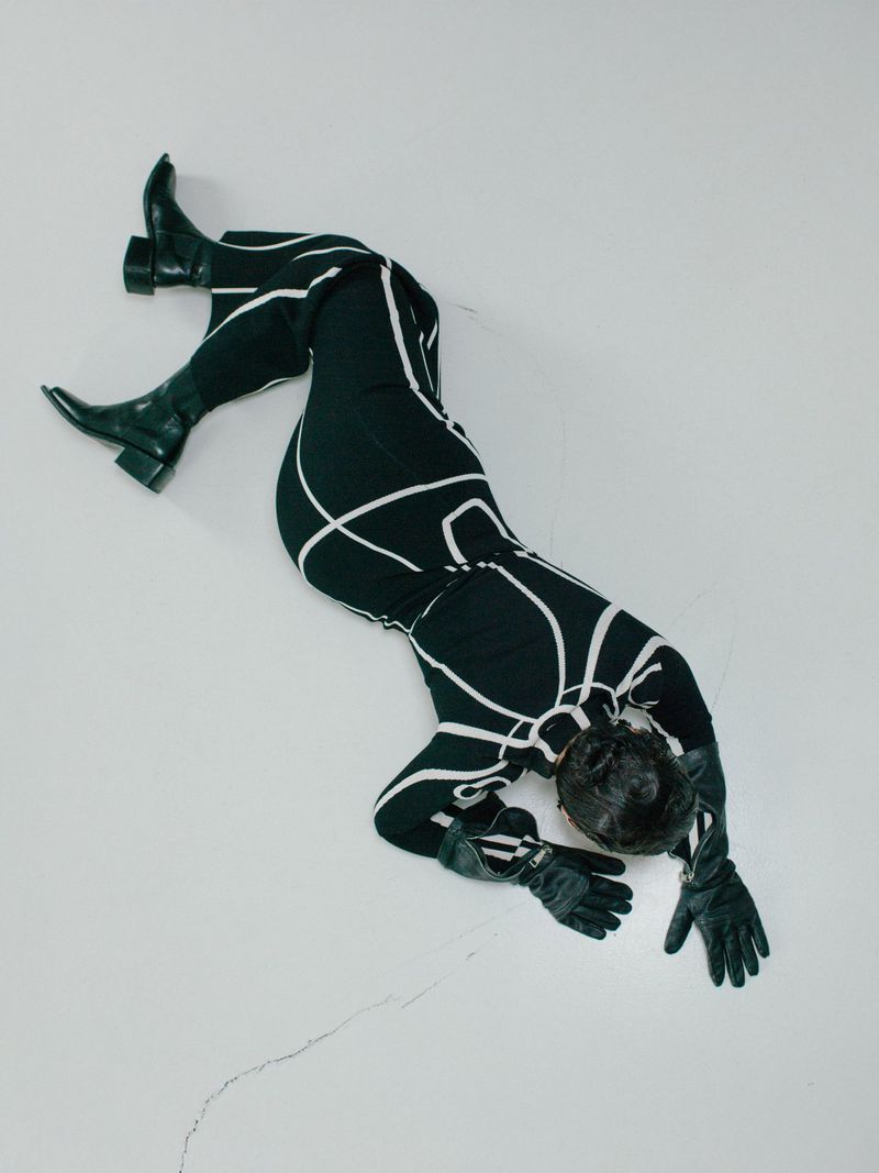 Overhead shot of a Black woman in a black and white catsuit and black boots on a white surface, showcasing fashion and style.