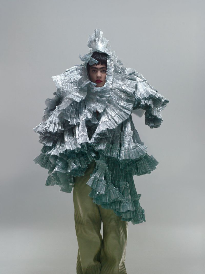 Person in avant-garde fashion wearing silver ruffled garment and green pants against a grey backdrop in a full shot.