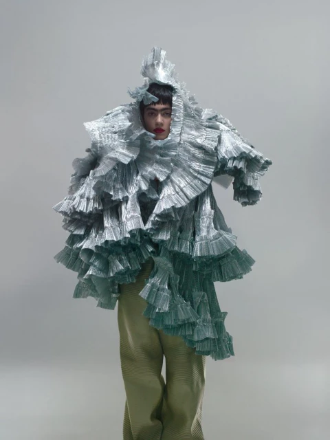 Person in avant-garde fashion wearing silver ruffled garment and green pants against a grey backdrop in a full shot.