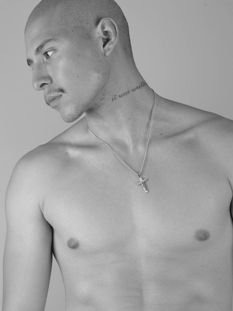 Monochrome portrait of a shirtless man wearing a silver cross necklace against a grey backdrop.