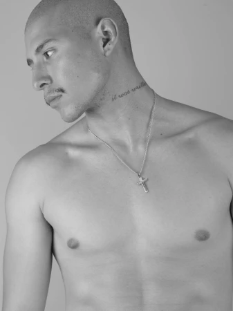 Monochrome portrait of a shirtless man wearing a silver cross necklace against a grey backdrop.