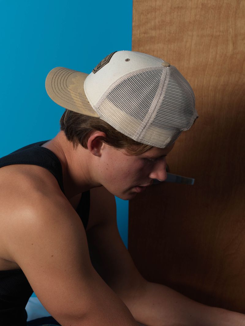 Young adult man wearing a baseball cap sitting in front of a blue and wood panel background in a medium indoor shot.