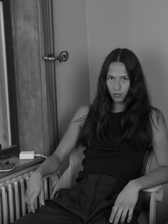 Portrait of a model with long dark hair wearing a black sleeveless top seated in a chair by a window.