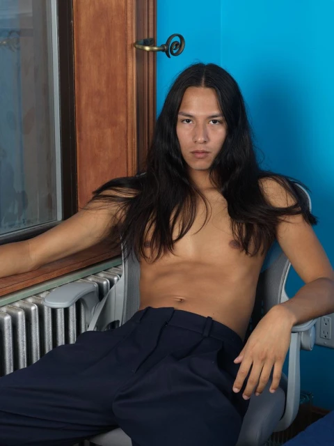 Portrait of a young man with long dark hair sitting shirtless in a chair in front of a blue wall and a window with a radiator.