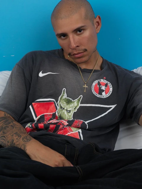Portrait of a bald adult male wearing a graphic tee, gold chain and black pants, seated against a blue wall.