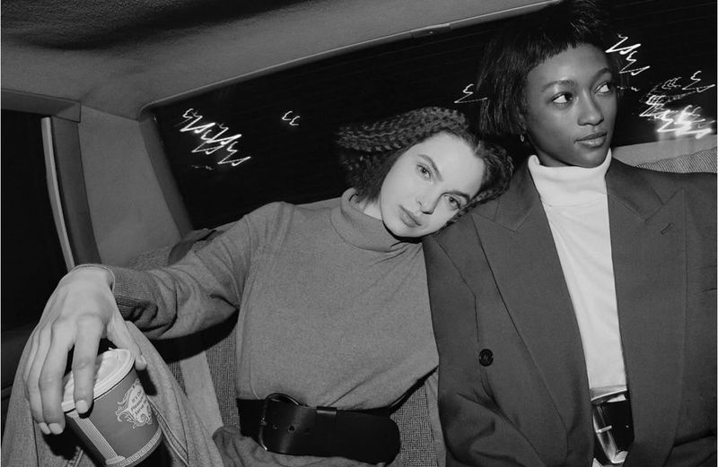 Two women pose in car interior with city lights at night. Woman in grey sweater rests head on blazer-clad friend as she holds coffee cup.