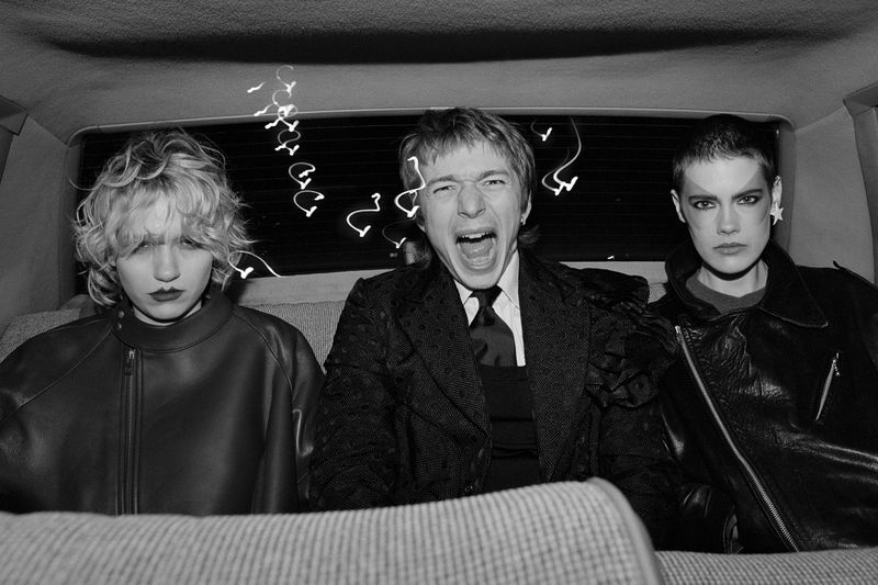 Black and white studio shot of three people in a car, one man yelling, two women staring at the camera, all wearing leather jackets.