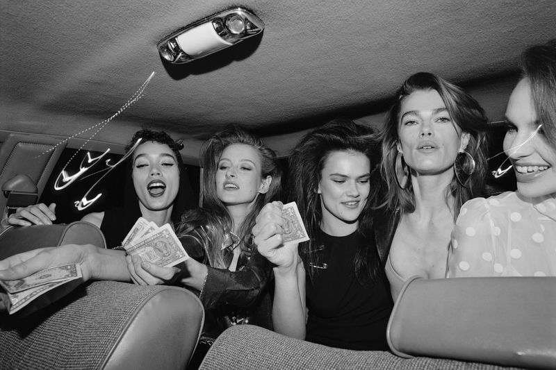 Five women are sitting in a car, holding money and wearing fashionable clothing, illuminated by the car's interior light in a black-and-white photograph.