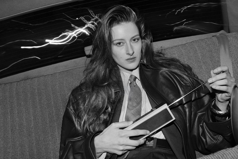 Black and white shot of a woman in a car, wearing a leather jacket, shirt, and tie, holding a device, with streaks of light visible.
