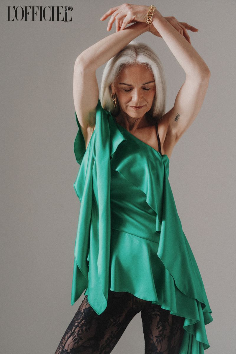 Mature woman in a draped green top and black lace pants poses with her arms up on a grey background for a fashion editorial shot.
