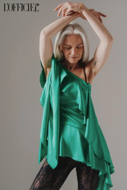 Mature woman in a draped green top and black lace pants poses with her arms up on a grey background for a fashion editorial shot.