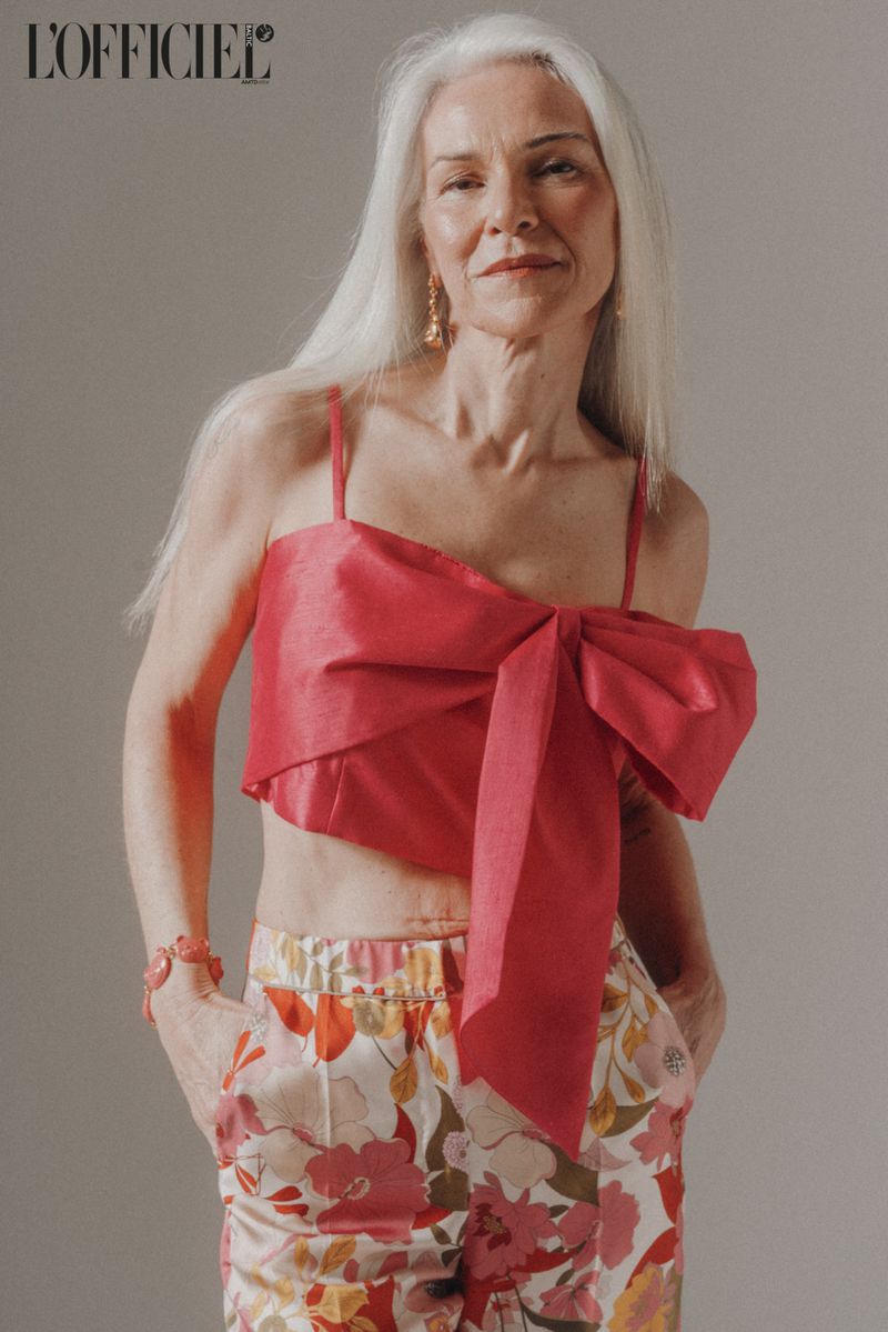 Fashion editorial shot of a mature woman with silver hair wearing a pink crop top with a bow and floral print pants.