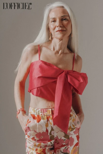 Fashion editorial shot of a mature woman with silver hair wearing a pink crop top with a bow and floral print pants.