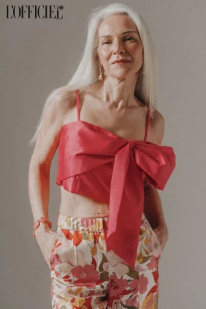 Fashion editorial shot of a mature woman with silver hair wearing a pink crop top with a bow and floral print pants.