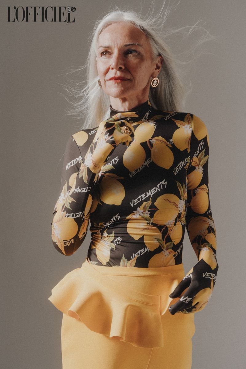 Elegant older woman poses in a black top with lemon print and a yellow skirt in a fashion editorial studio shot. 